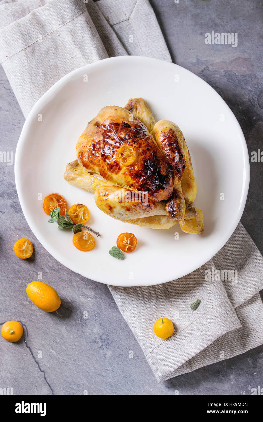 Whole roasted mini chicken cooking with caramelized kumquats served on white plate with textile napkin and fresh citrus fruits over gray kitchen table Stock Photo