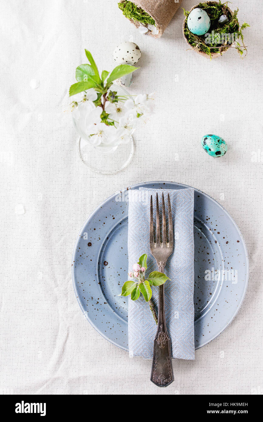 Table setting decor colorful Easter quail eggs with spring cherry flowers, moss in garden pots, empty plates, vintage cutlery over white tablecloth. T Stock Photo