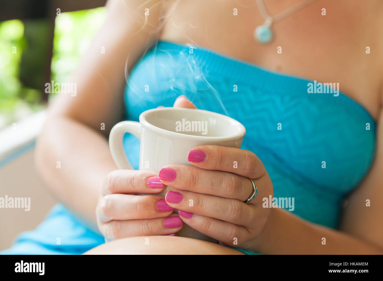 Young Caucasian woman in blue dress holds white cup of coffee in her hands. Close-up outdoor photo, selective focus on hands Stock Photo