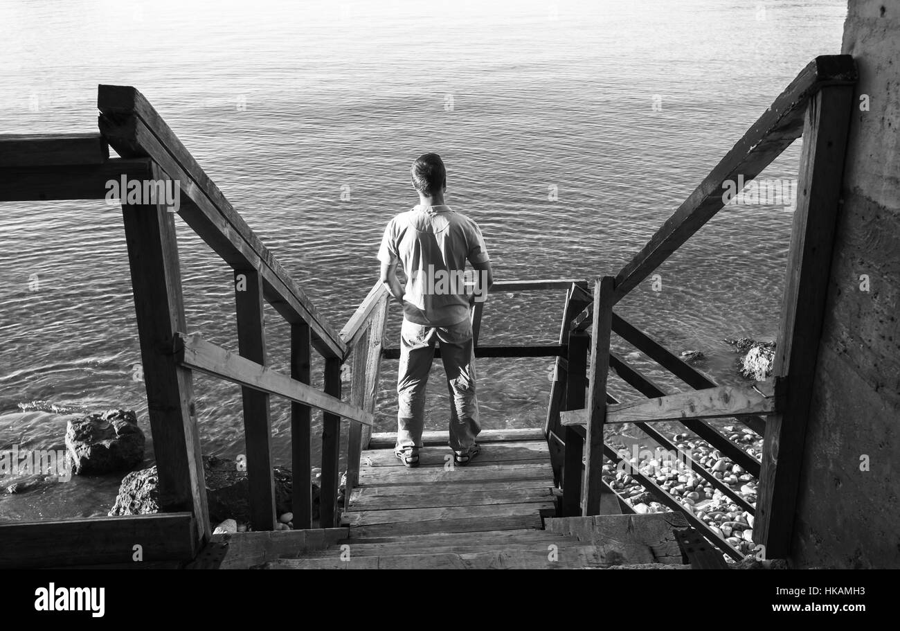 Young man stands on old wooden stairway going down to the sea coast, black and white photo Stock Photo