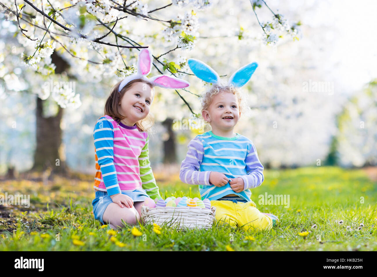 Kids on Easter egg hunt in blooming garden. Children with bunny ears searching for colorful eggs in snow drop flower meadow Stock Photo