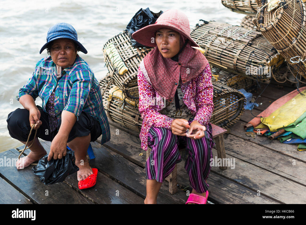 Two women sit on a wooden dock at the water's edge with a pile of crab traps behind them, local life in the famous crab market; Kep, Cambodia Stock Photo