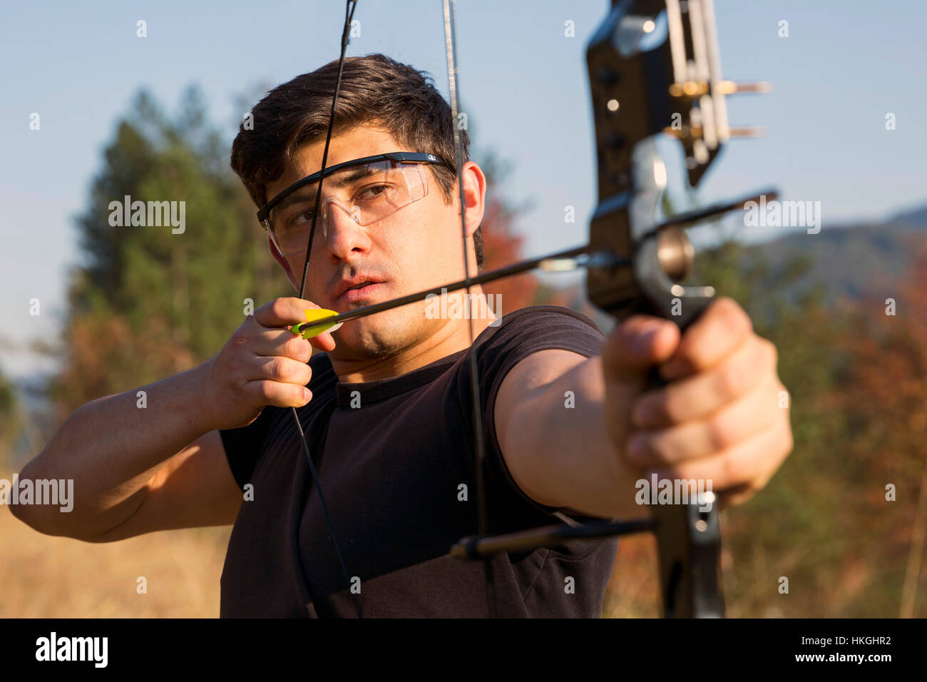 An archer draws his compound bow and aims upwards with clean blue sky as background. Stock Photo