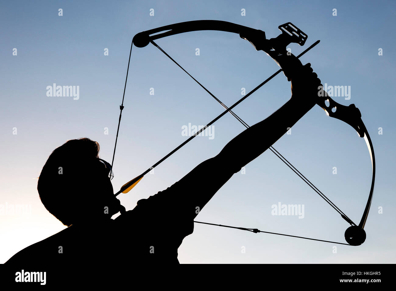 A silhouette of an archer draws his compound bow and aims upwards with clean blue sky as background. Stock Photo