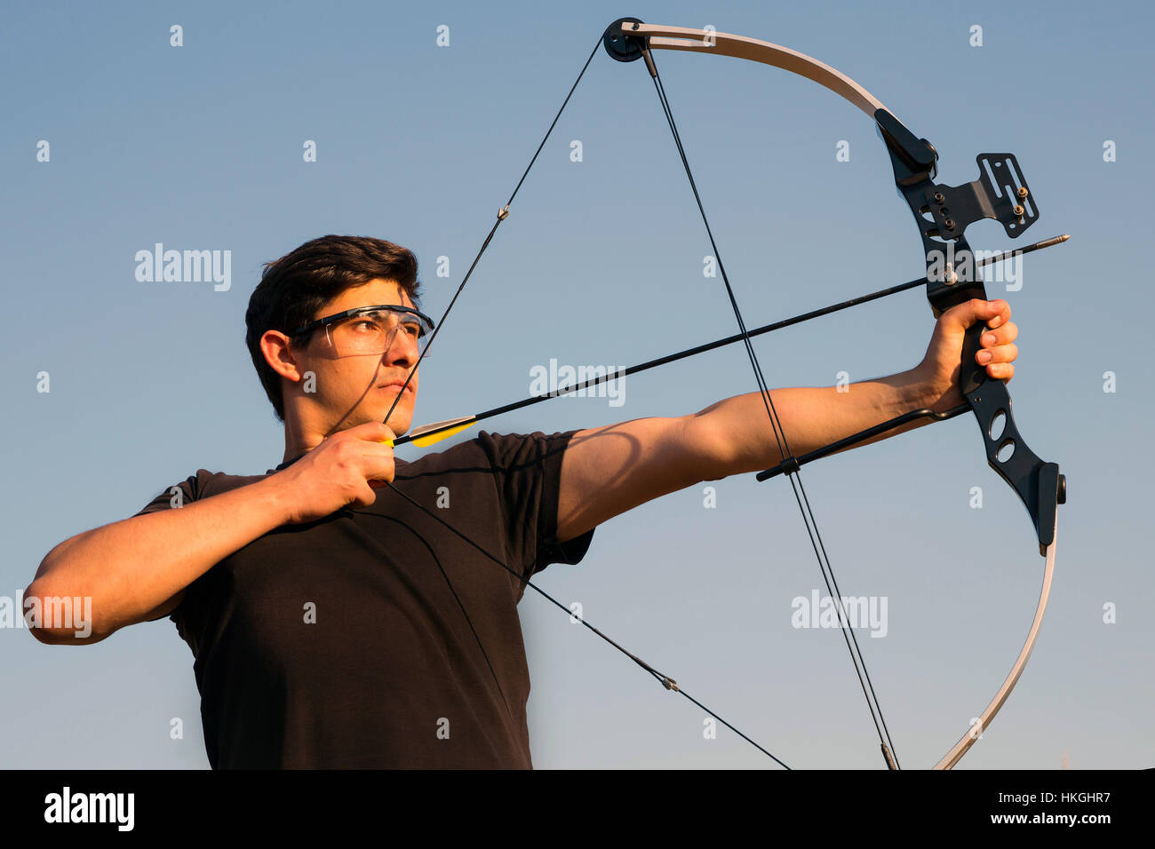 An archer draws his compound bow and aims upwards with clean blue sky as background. Stock Photo