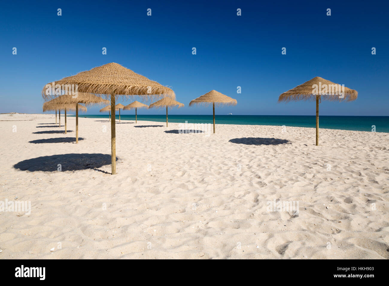 Straw umbrellas on empty white sand beach with clear sea behind, Ilha do Farol, Culatra barrier island, Olhao, Algarve, Portugal Stock Photo