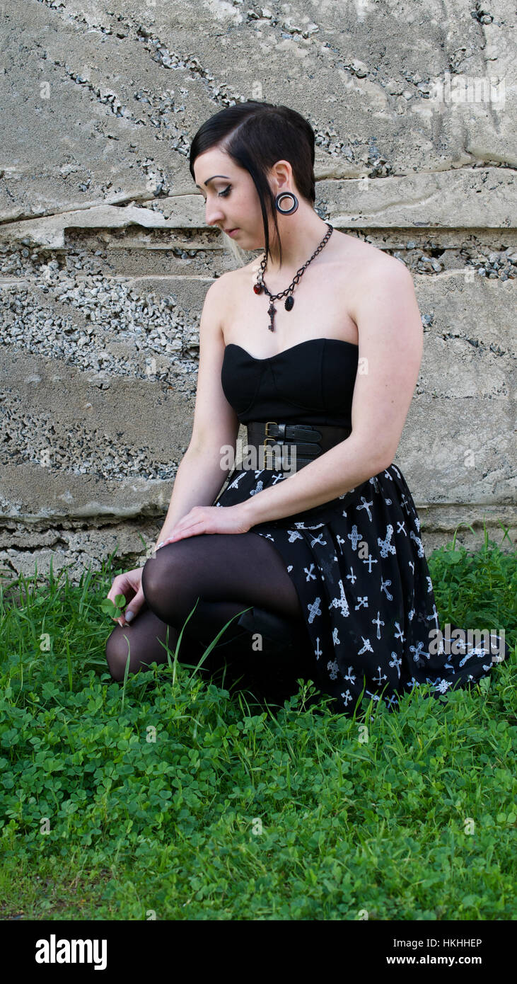 Beautiful, young gothic brunet female model kneeling down on the grassand, handling a blade of grass, against cement wall - Stock Photo