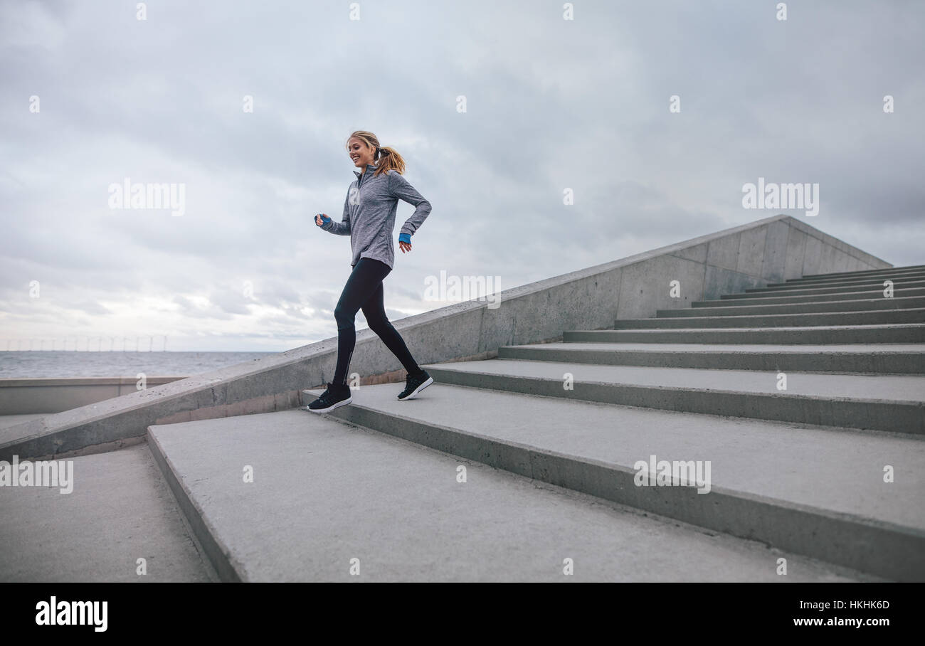 Full length shot of healthy young woman running down on steps and smiling. Female runner exercising on stairs in morning. Stock Photo