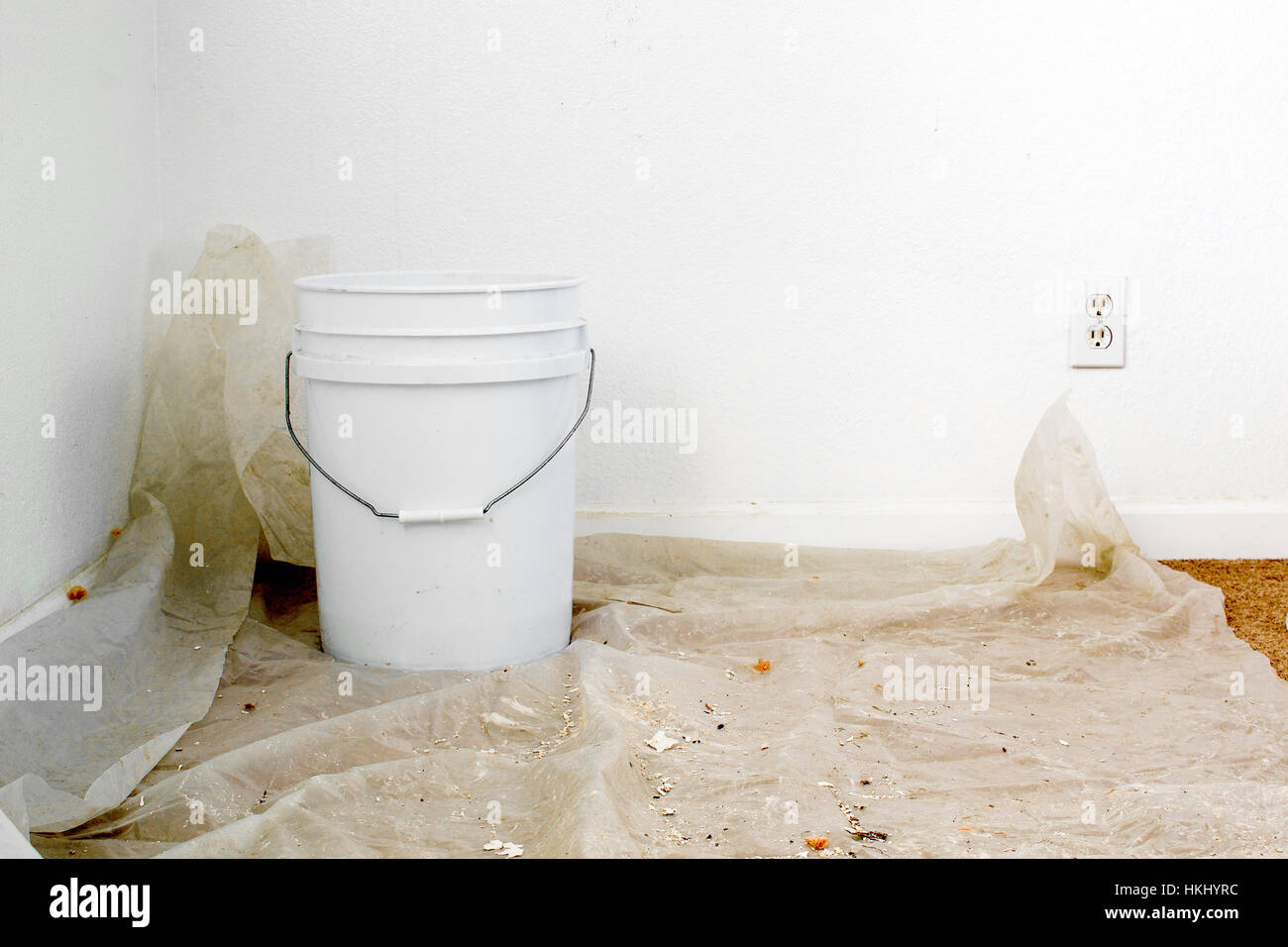 A white bucket over a sheet of plastic placed over carpet floor to collect water from leaking ceiling Stock Photo
