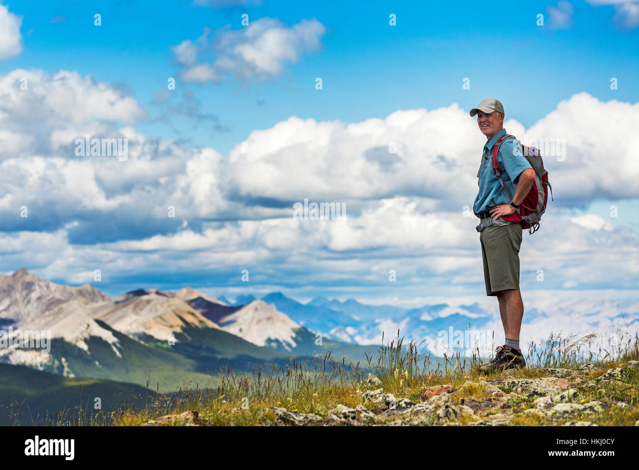 Male hiker standing on top of rocky hill overlooking mountain range and valley with blue sky and clouds, West of Bragg Creek; Alberta, Canada Stock Photo