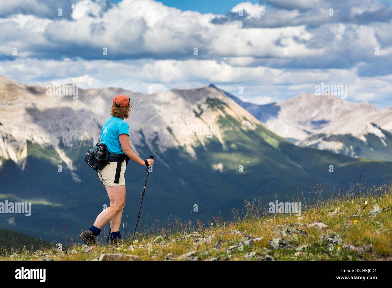 Female hiker walking along a rocky hill overlooking mountain range and valley with a cloudy sky, West of Bragg Creek; Alberta, Canada Stock Photo