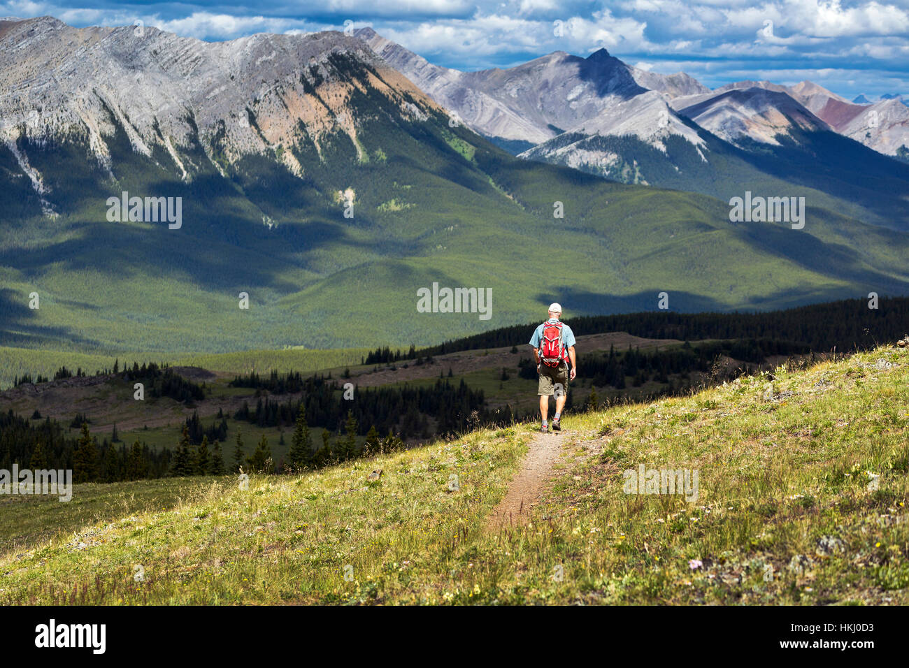 Male hiker walking along a hillside trail overlooking mountain range and valley with a cloudy sky, West of Bragg Creek; Alberta, Canada Stock Photo
