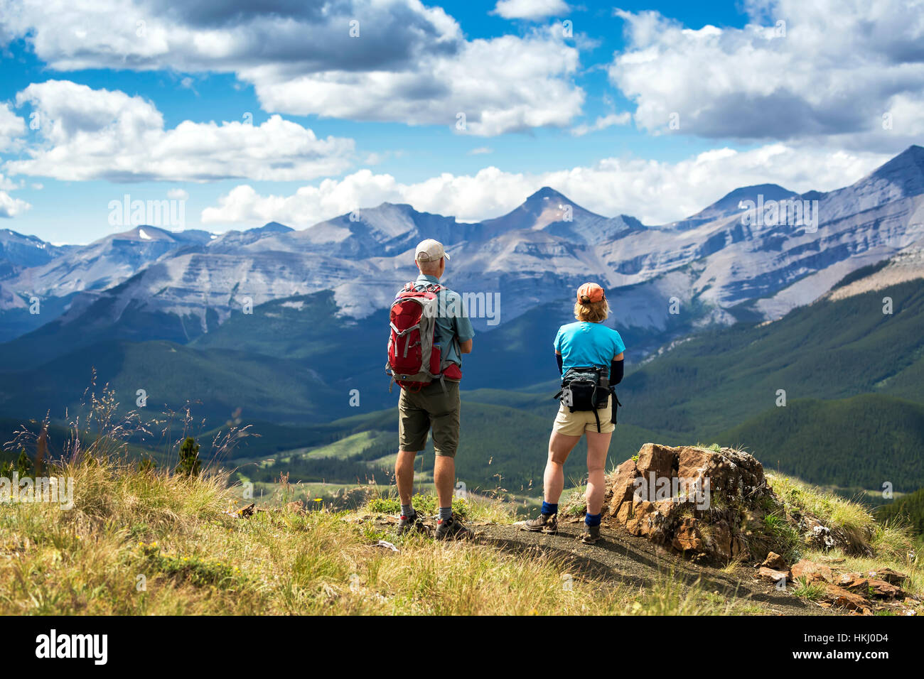 Male and female hikers standing on top of rocky hill overlooking mountain range and valley with blue sky and clouds, West of Bragg Creek Stock Photo