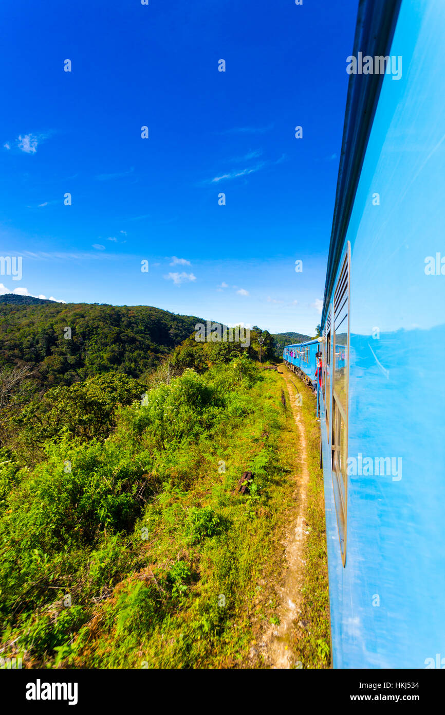 Exterior view out the side of a moving blue passenger train in hill country landscape on a blue sky day near Newara Elia Stock Photo