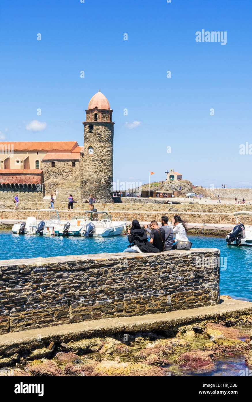 People seated on a wall looking towards the Church of Notre Dame des Anges and Chapelle Saint-Vincent, Collioure, Côte Vermeille, France Stock Photo