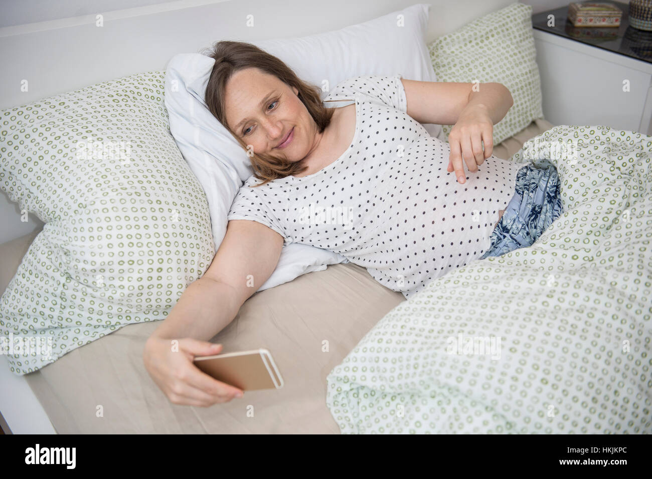 Pregnant woman lying in bed and taking selfie on mobile phone, Munich, Bavaria, Germany Stock Photo