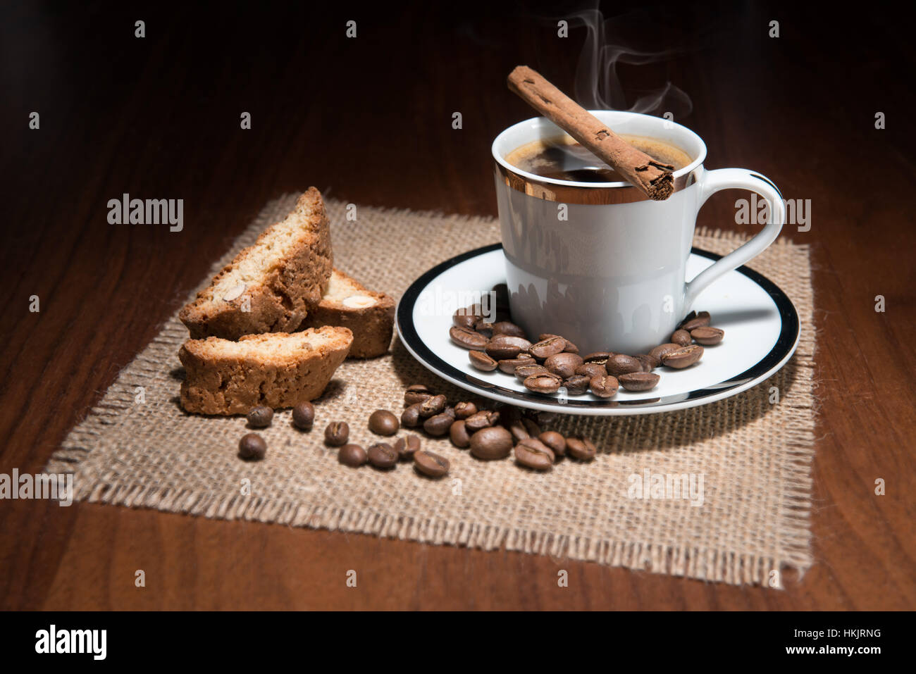 A studio, still life of a cup of coffee on a saucer with a cinnamon stick, roasted beans and biscuits on a hessian mat Stock Photo