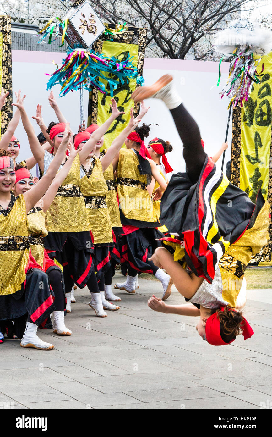 Japanese Yosakoi dance festival. Young women team, in black, gold and red costume, formation dancing in city square. Dancer somersaulting. Stock Photo