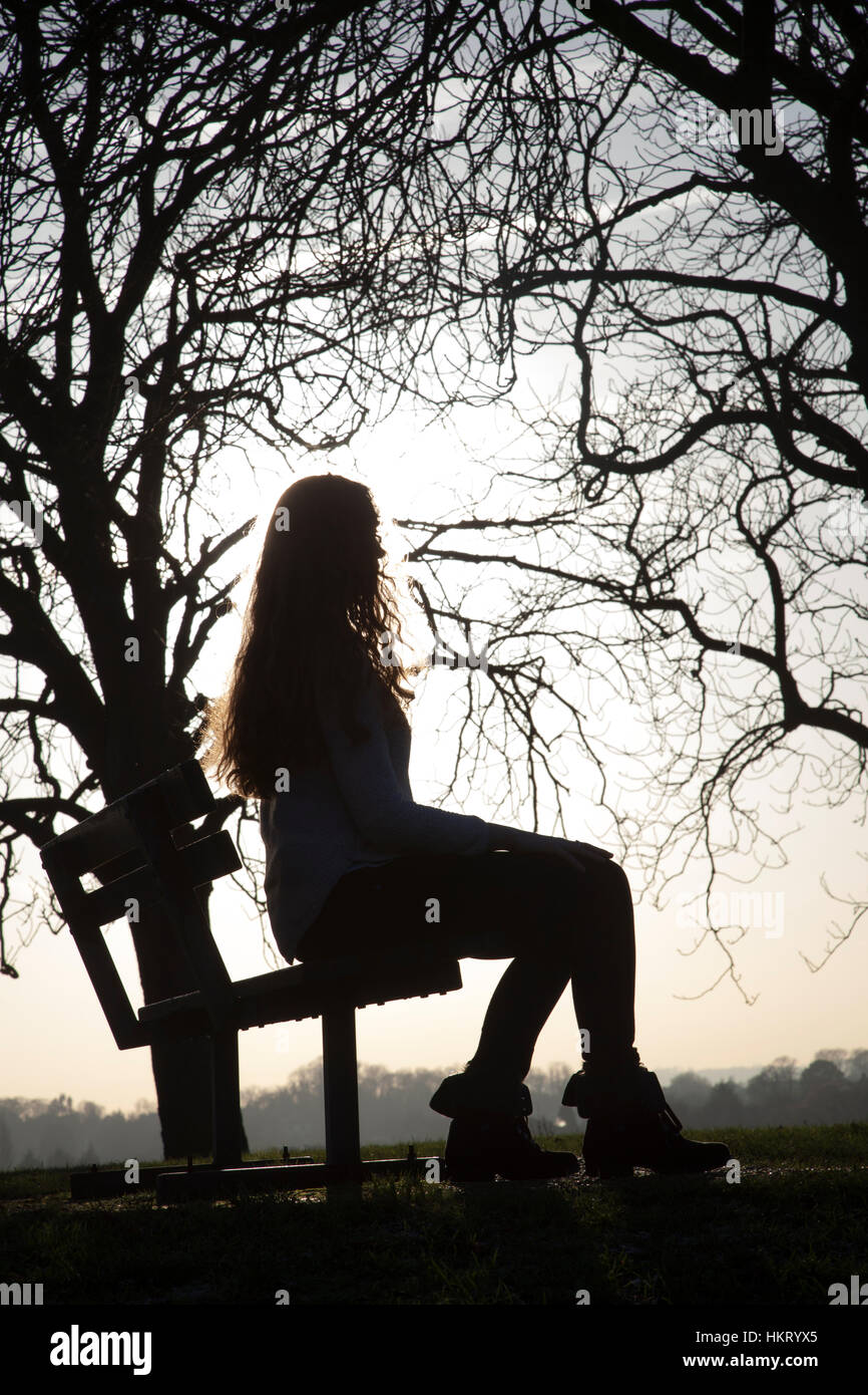 Young pensive woman in 20s with long hair, sitting on a park bench looking into the distance, outdoors, silhouette. Stock Photo