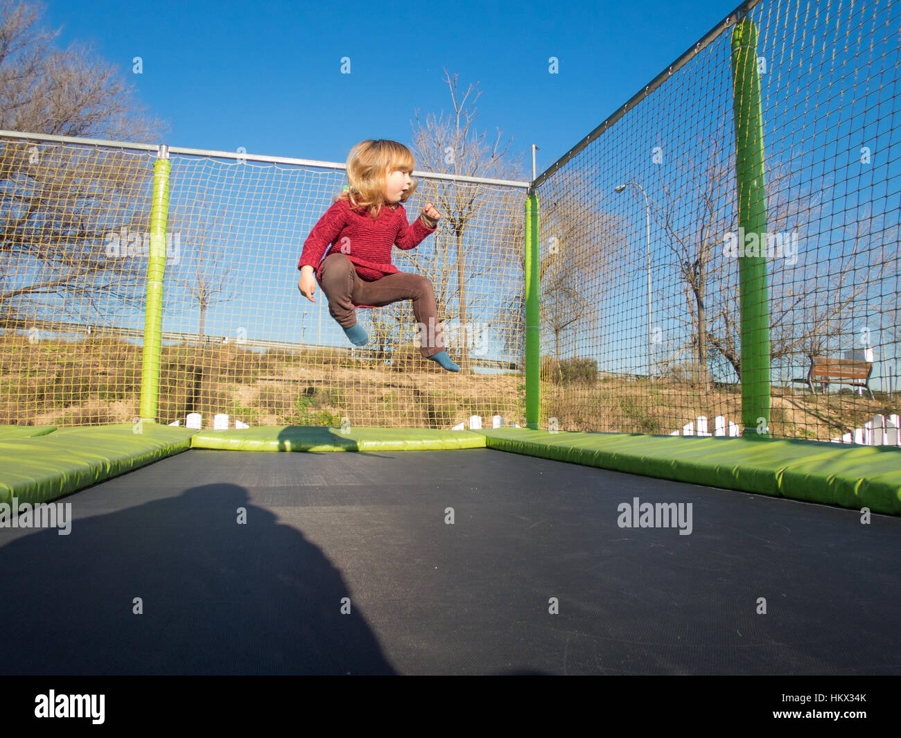 Happy three years old blonde child with winter red sweater trampolining or jumping on trampoline in outdoor playground Stock Photo