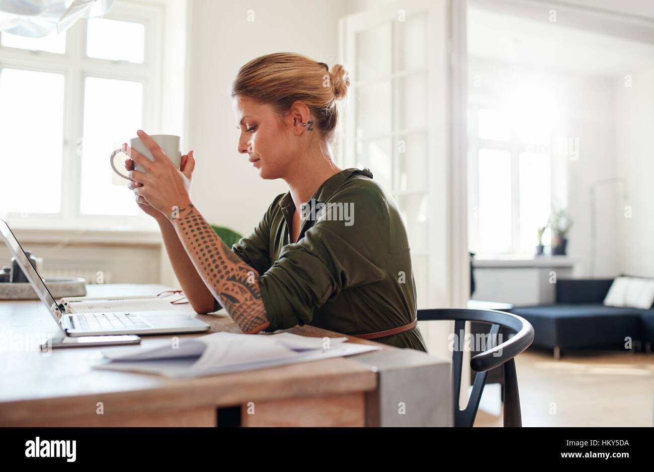 Side view shot of thoughtful young woman with coffee sitting at table. Female sitting at home office looking serious. Stock Photo