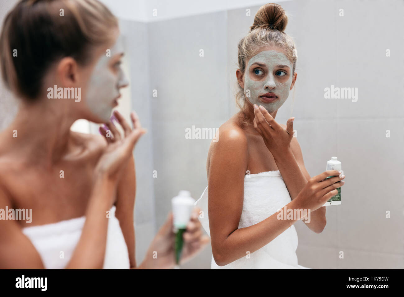 Young woman applying mask cream on face in bathroom. Female in doing beauty treatment on her face skin in front of mirror after bath. Stock Photo