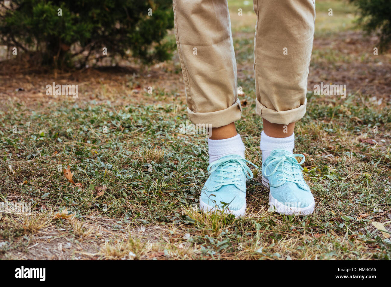 Female feet in beige pants and a turquoise sneakers standing on the grass near the trees. Autumn Walk. Stock Photo