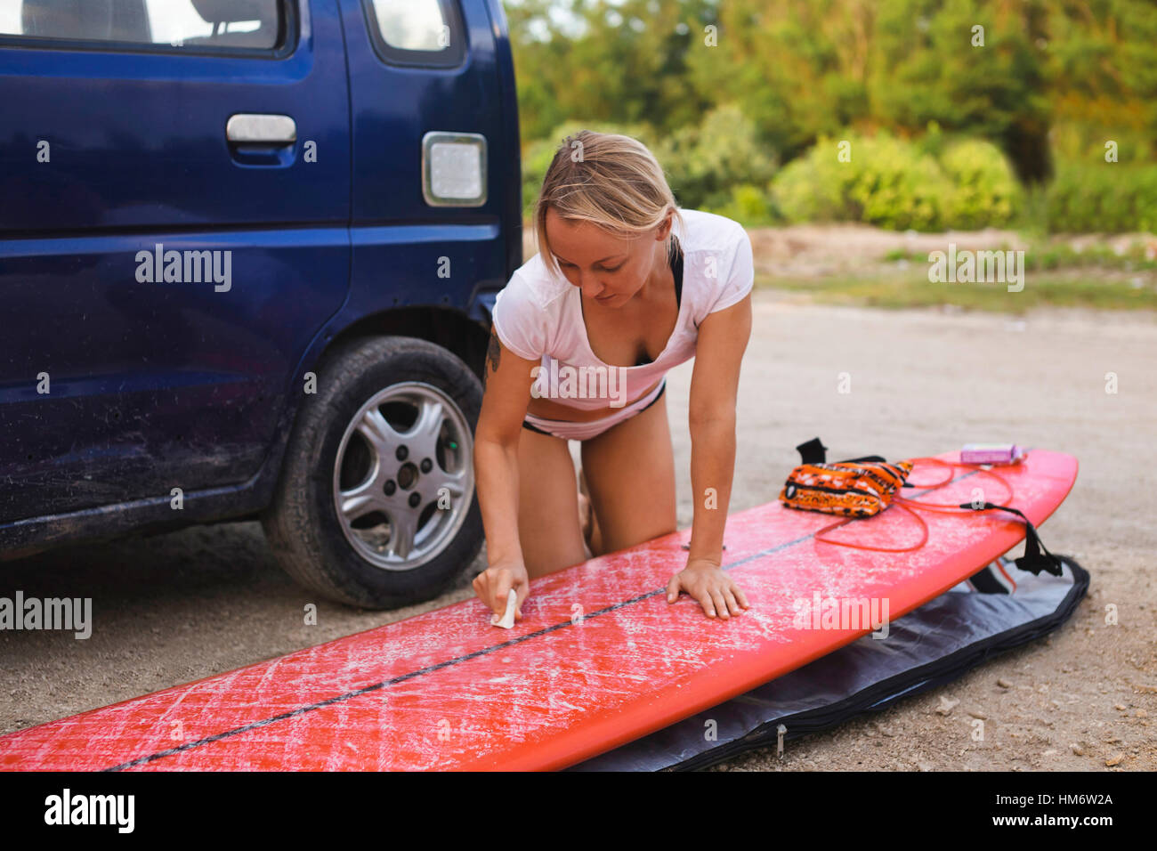 Woman waxing surfboard while kneeling by car on field Stock Photo