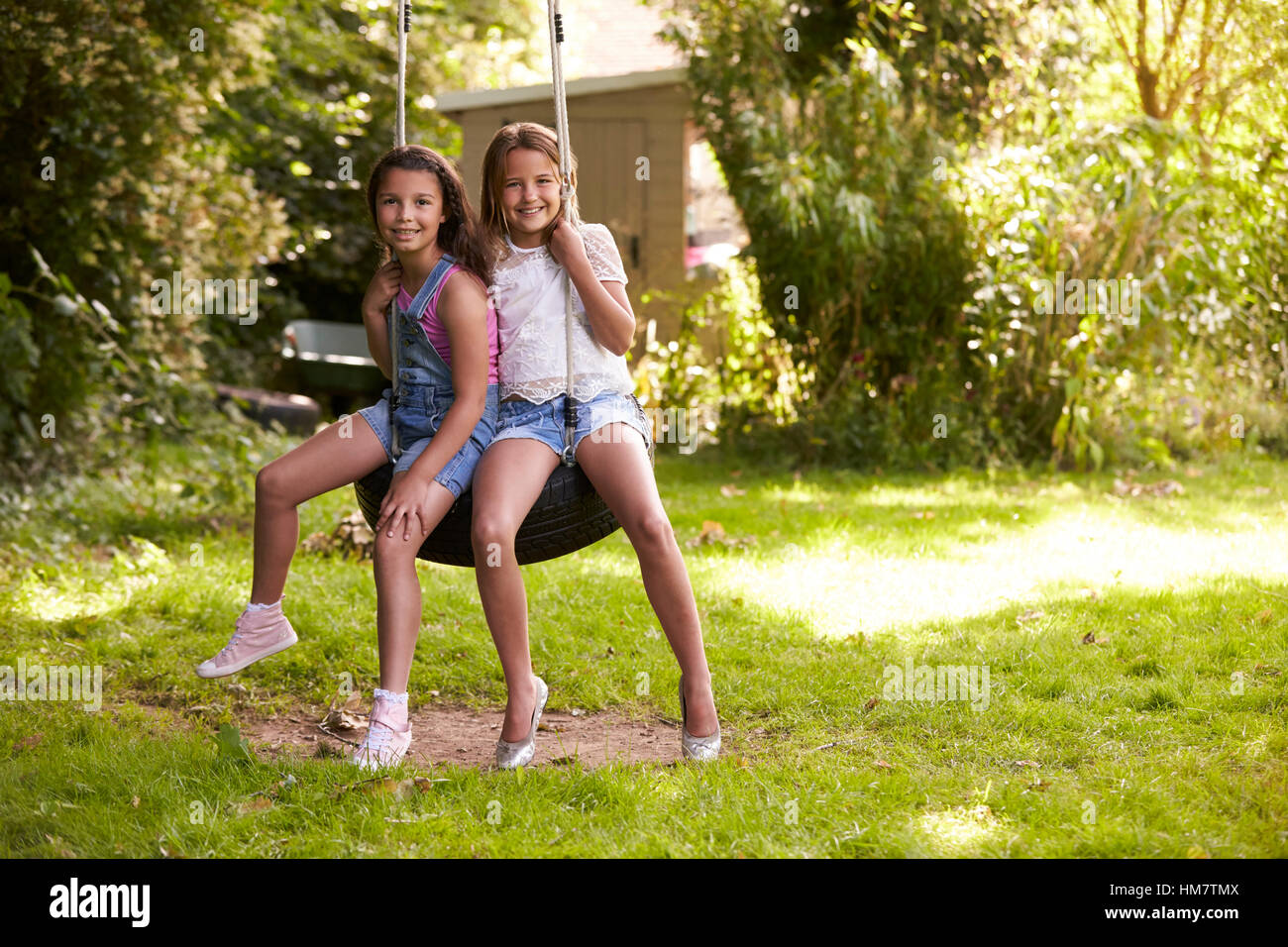 Portrait Of Two Girls Playing On Tire Swing In Garden Stock Photo