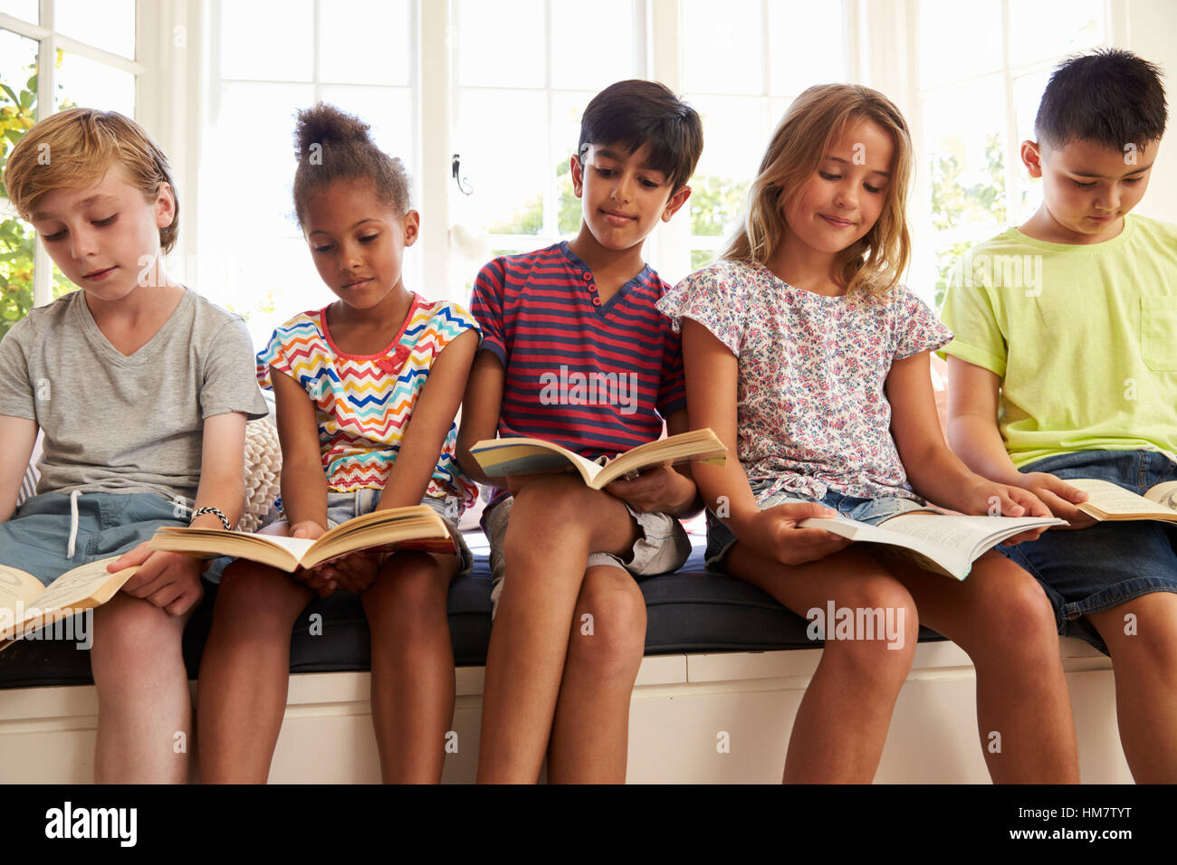 Group Of Multi-Cultural Children Reading On Window Seat Stock Photo