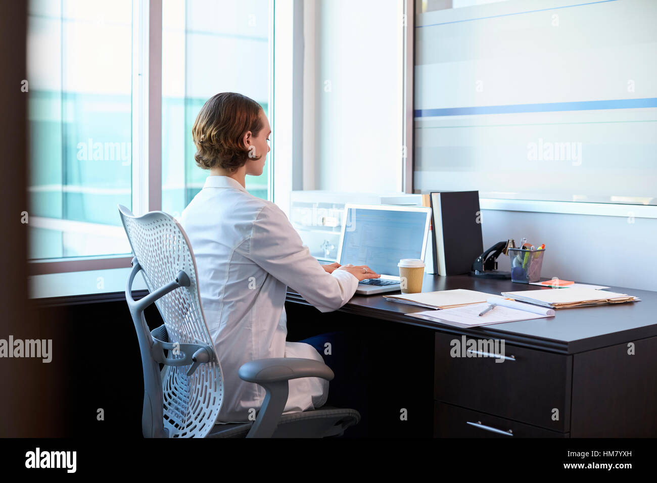 Female Doctor Wearing White Coat In Office Working At Desk Stock Photo