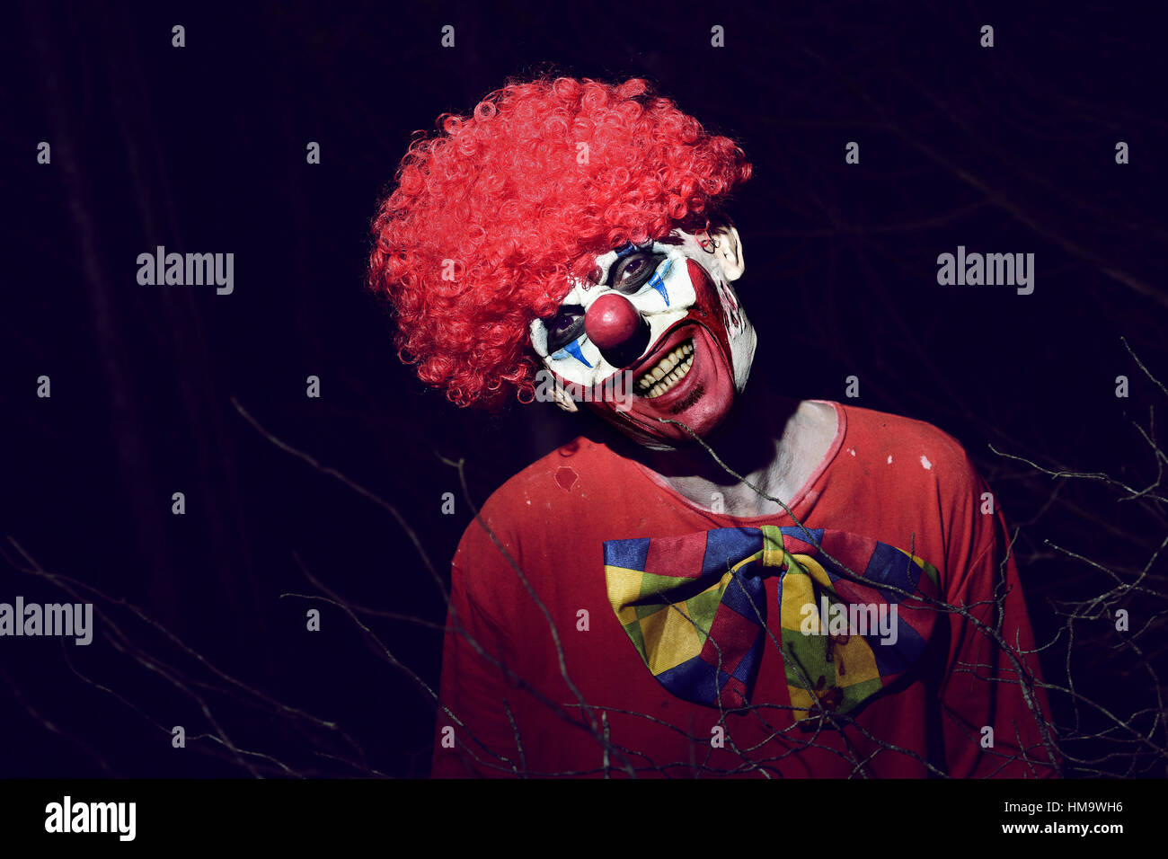 closeup of a scary evil clown wearing a red wig and a dirty costume, in the woods at night Stock Photo