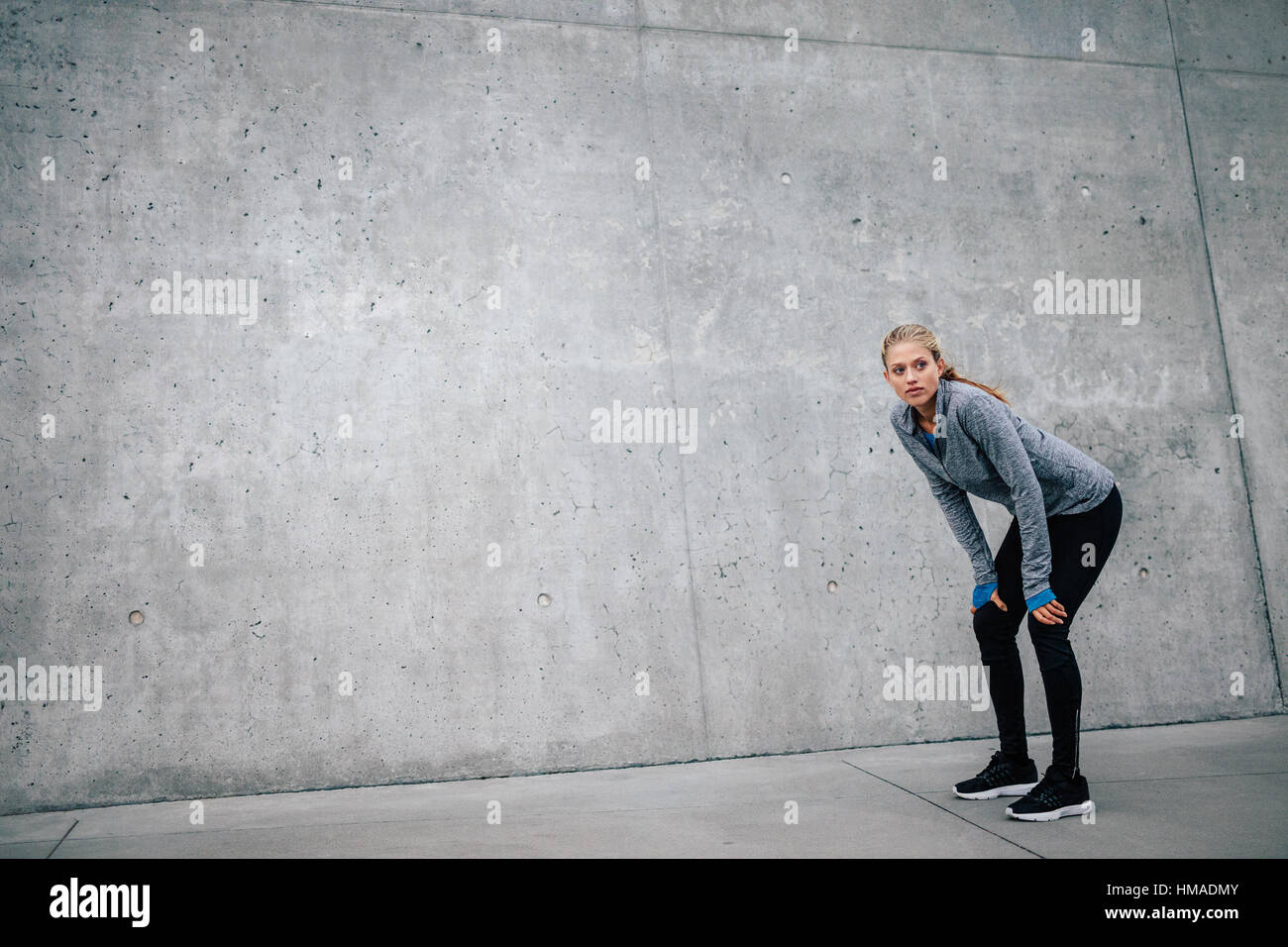 Full length shot of young beautiful female leaning on gray background while resting after everyday training. Woman athlete runner taking a break with  Stock Photo
