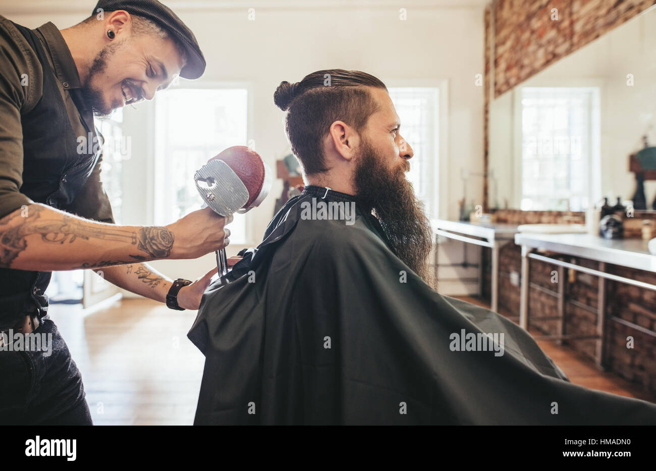 Handsome bearded man in a salon cape in the barbershop, with hairdresser adjusting the headrest of the chair. Stock Photo