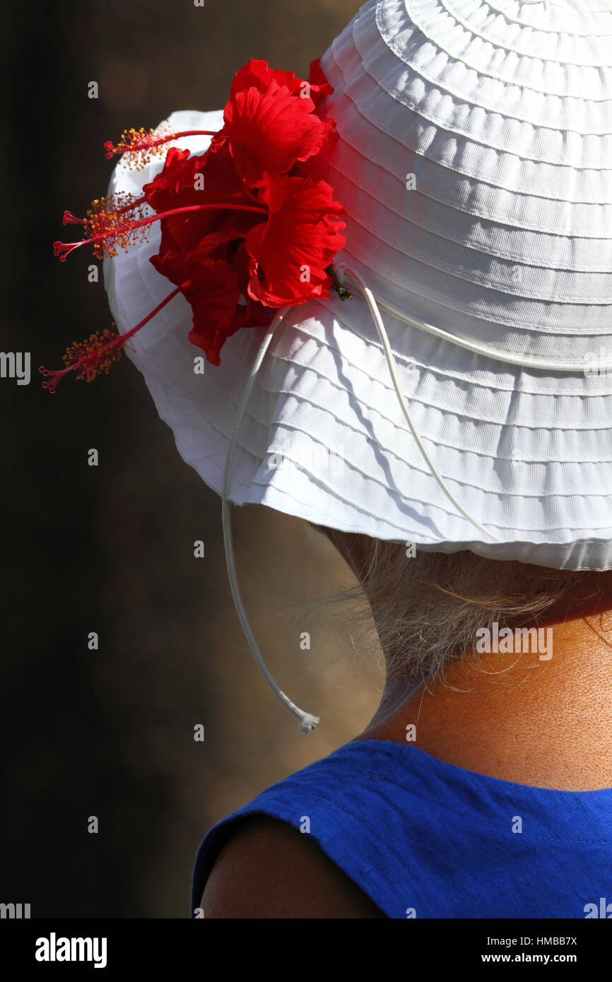 A mature lady in blue singlet wears a white hat with red hibiscus flower during summer in Queensland, Australia. Stock Photo