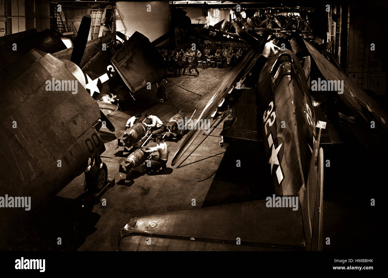 Surrounded by F6F's, ordnancemen work on bombs on hangar deck of USS YORKTOWN.  Officers and men in background watch movie.  Ca.  1943.  Lt. Comdr. Charles Kerlee.  (Navy) Stock Photo
