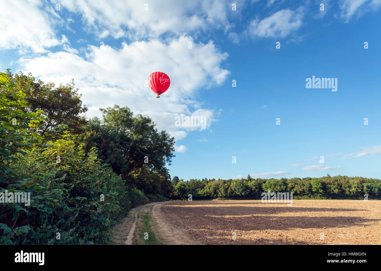 Virgin hot air balloon drifts over newly ploughed farm field and trees in the Chilterns on a summer evening Stock Photo