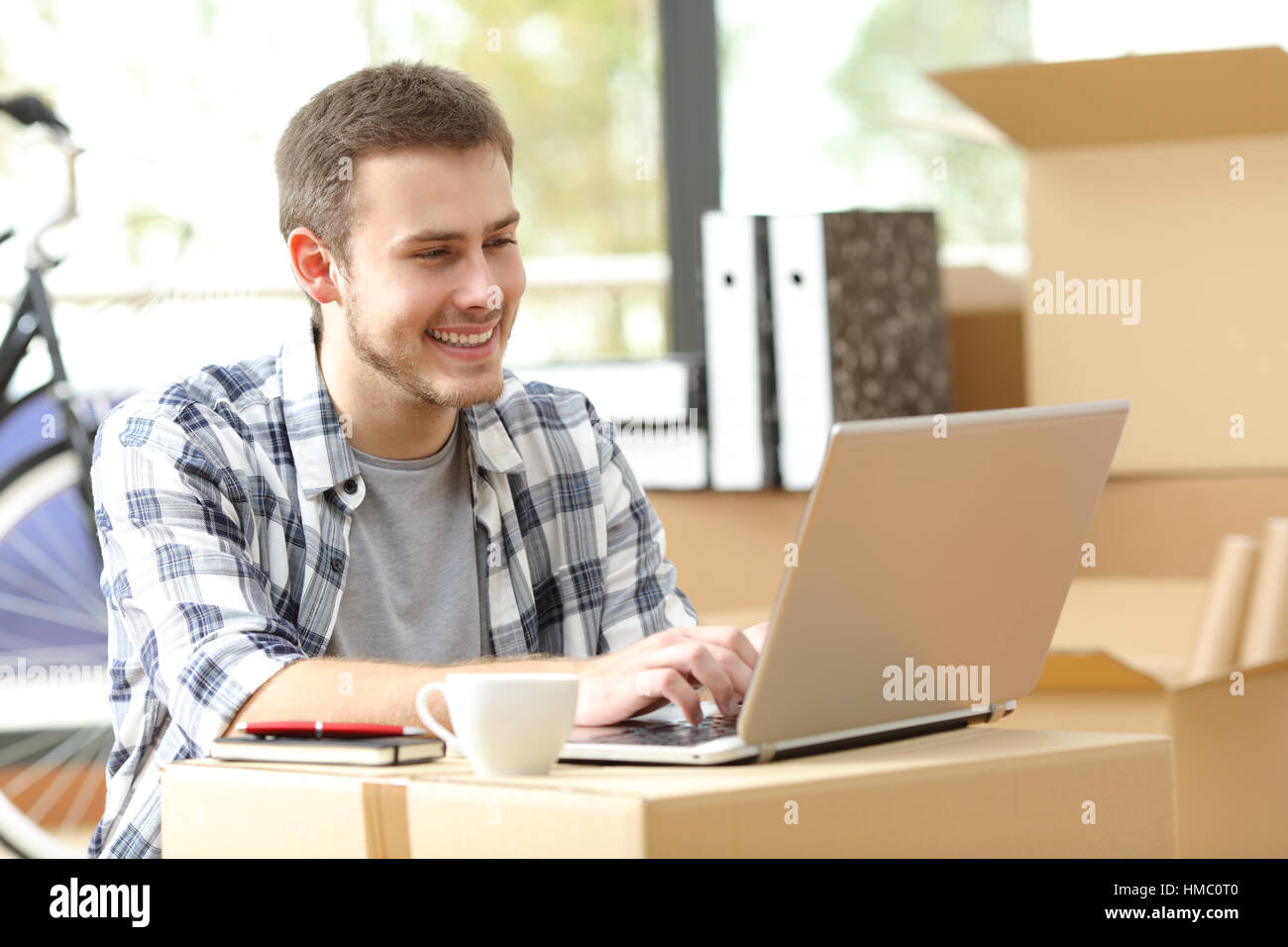 Happy entrepreneur sitting on the floor working on line in his new workplace using a carton box like a desktop while is moving office Stock Photo