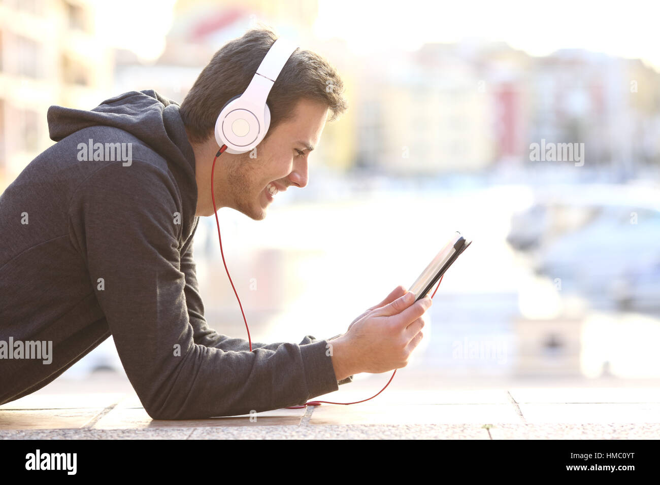 Side view of a happy teenager listening music or watching videos on line in  a tablet lying on the floor with a port in the background Stock Photo -  Alamy