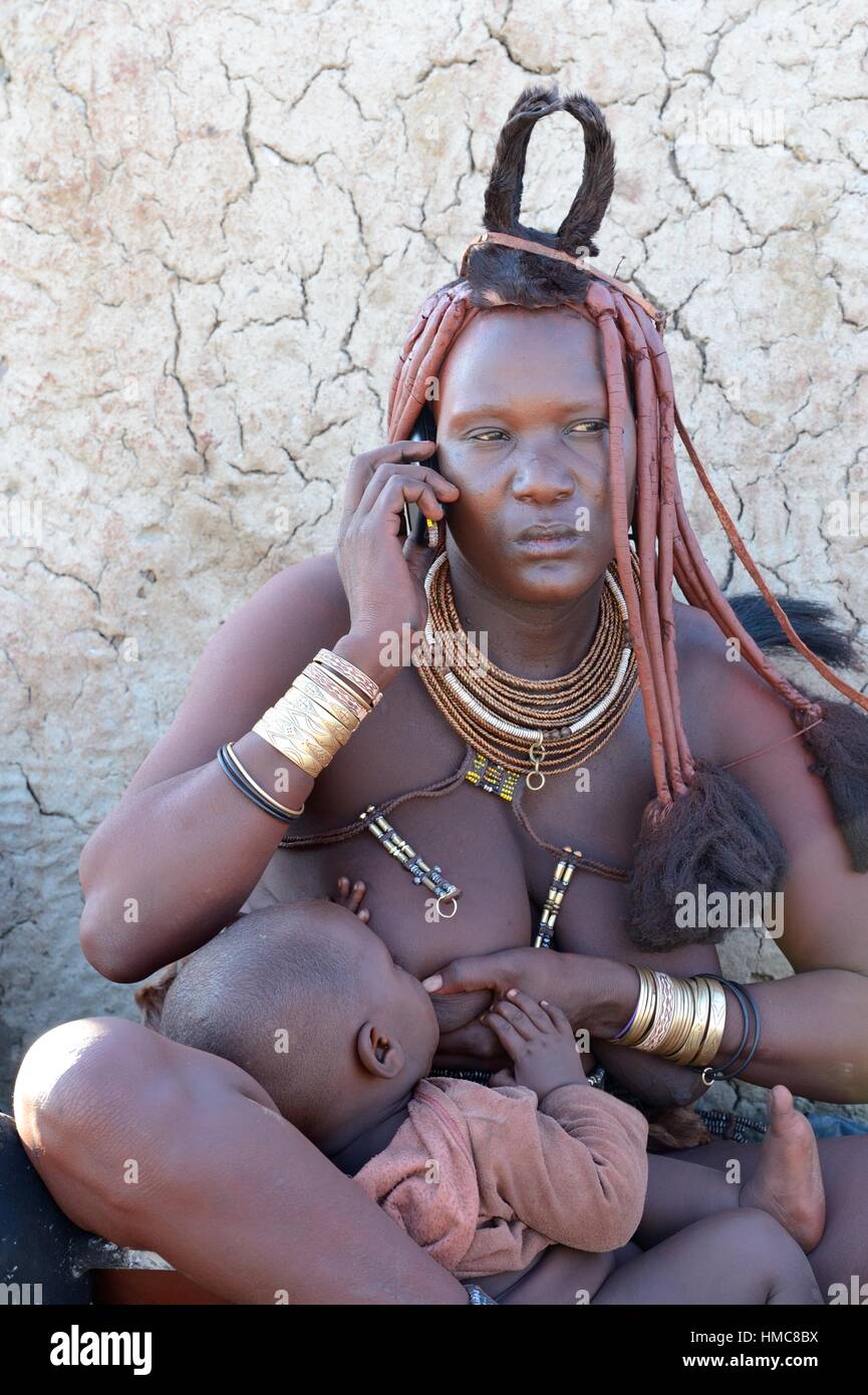 Himba woman with the typical ornaments and a cell phone nursing her baby. Kaokoland, Namibia. Stock Photo