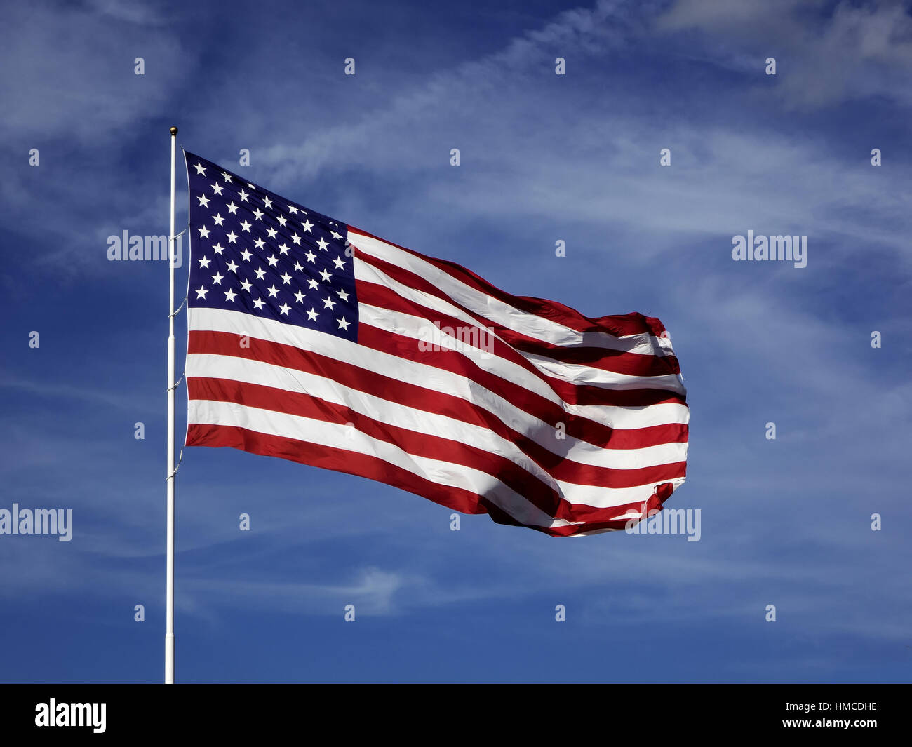 An oversized American flag raised on a flagpole waves in the wind against a blue sky background with white clouds. Stock Photo