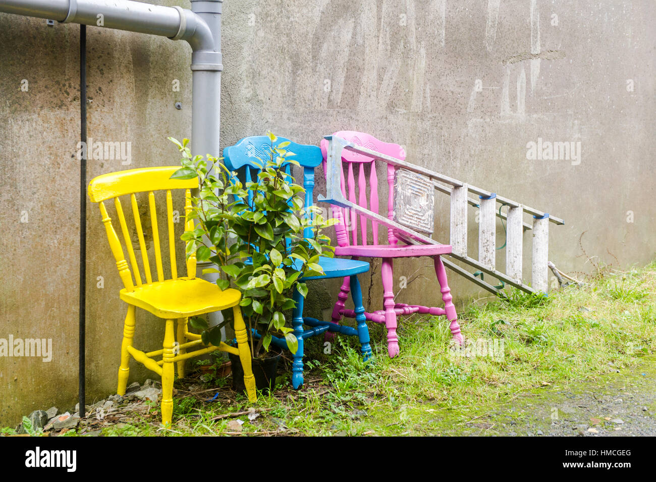 Three recycled/upcycled colourful chairs and a step ladder against a wall in an alleyway with grass and copy space. Stock Photo