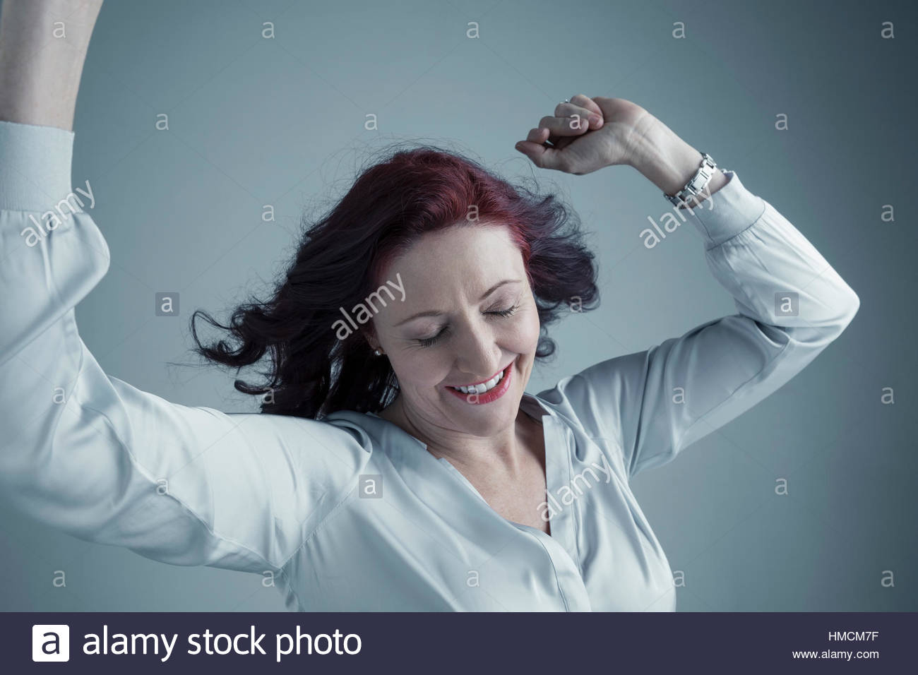 Portrait carefree Caucasian mature woman with burgundy red hair dancing with arms raised and eyes closed Stock Photo