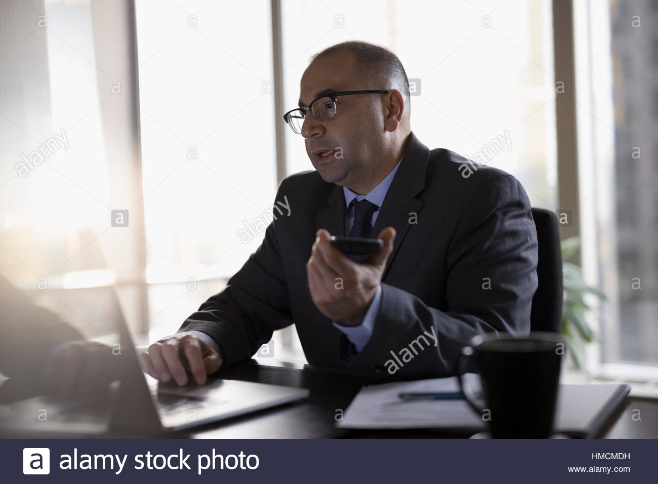 Male lawyer at laptop dictating notes with smart phone in office Stock