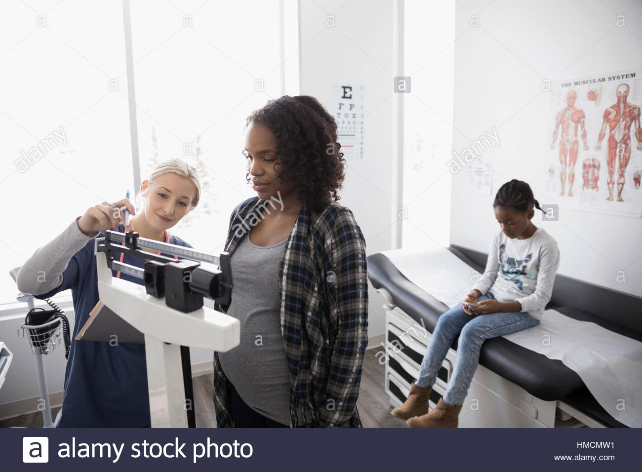 Female nurse checking weight of pregnant patient on scale in clinic examination room Stock Photo