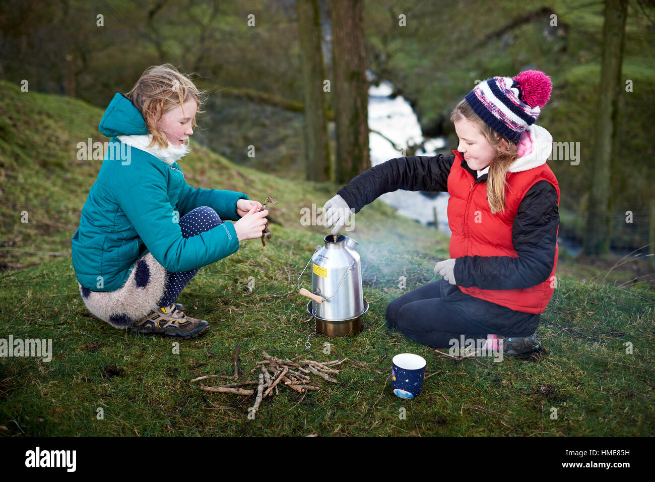 Two girls feed wood fuel into a Kelly Kettle solid fuel strove in woodland during winter Stock