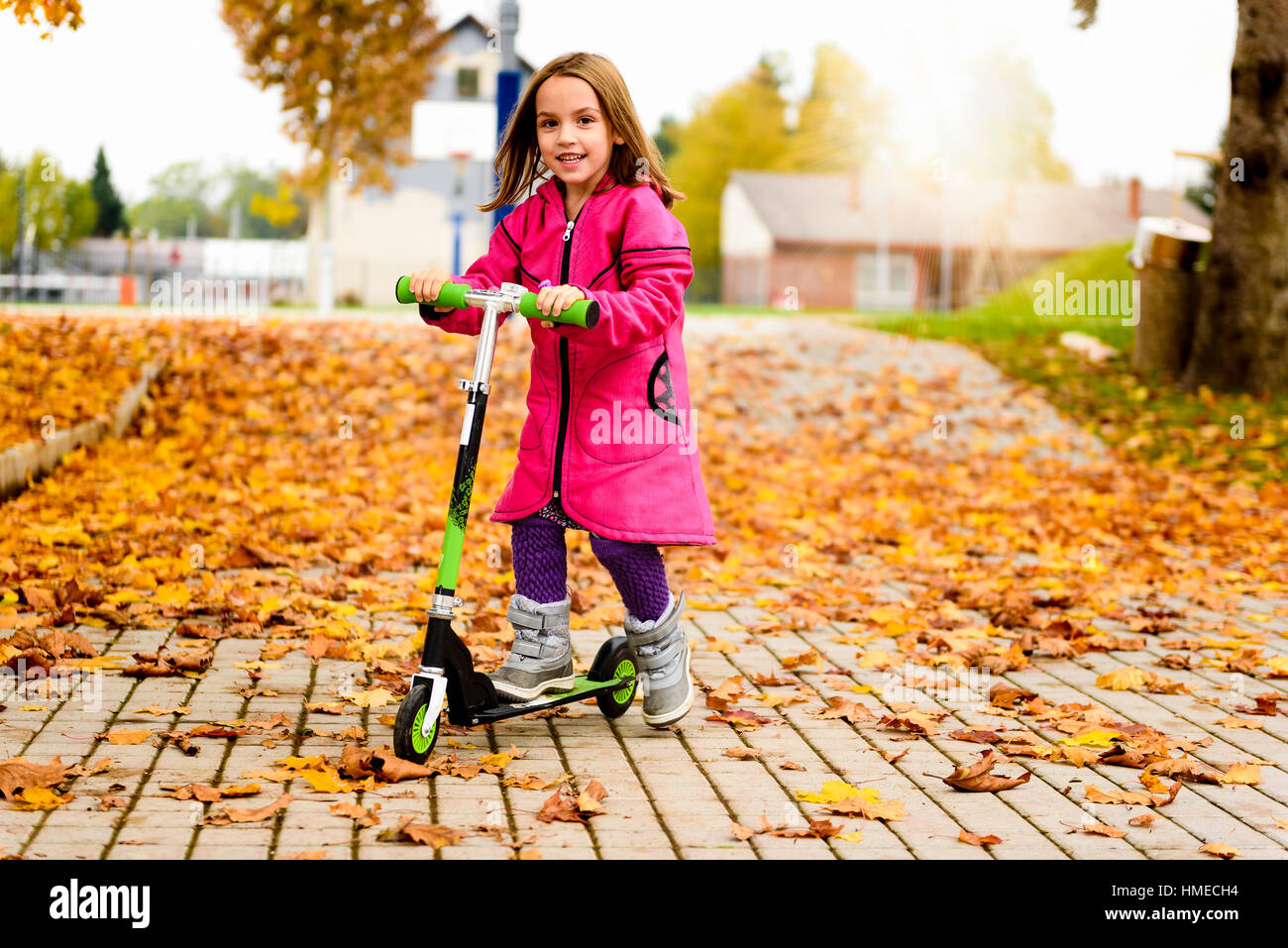 Girl in pink coat is riding scooter on maple leaves. Active child in sport pursuit is exercising in the nature in the autumn with cold temperatures ou Stock Photo