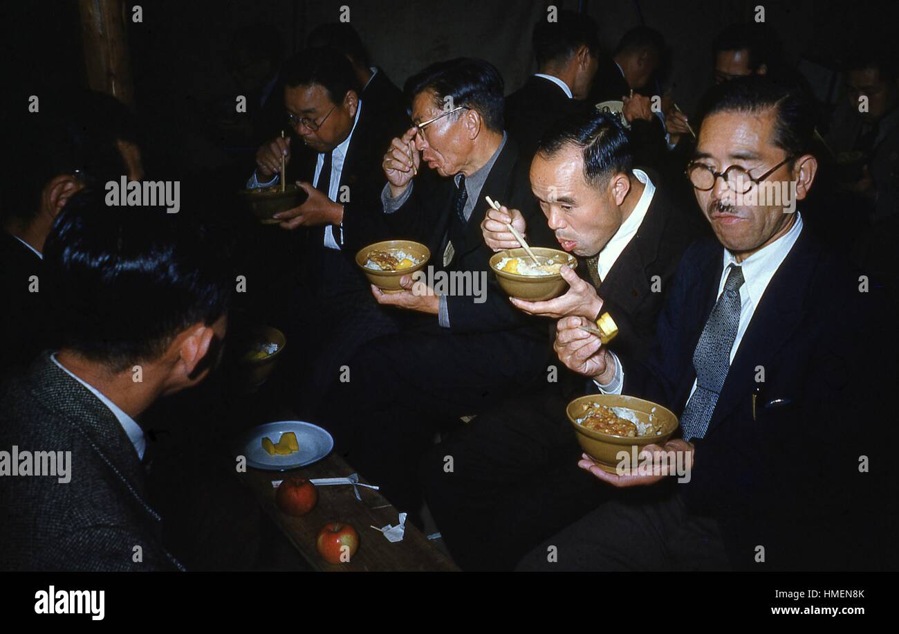 Japanese men eating ramen, rice, and eggs with chopsticks, dressed in formal suits, wearing glasses, with apples and other food on a table, in a crowded room, Japan, 1952. Photo credit Smith Collection/Gado/Getty Images. Stock Photo