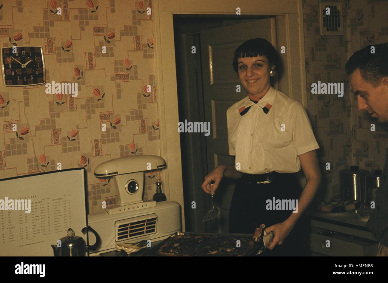 Woman standing in a 1950s kitchen holding a tray of food and a spatula over the stove, a coffee percolator can be seen at the bottom left, a man looking at the tray of food, 1952. Stock Photo