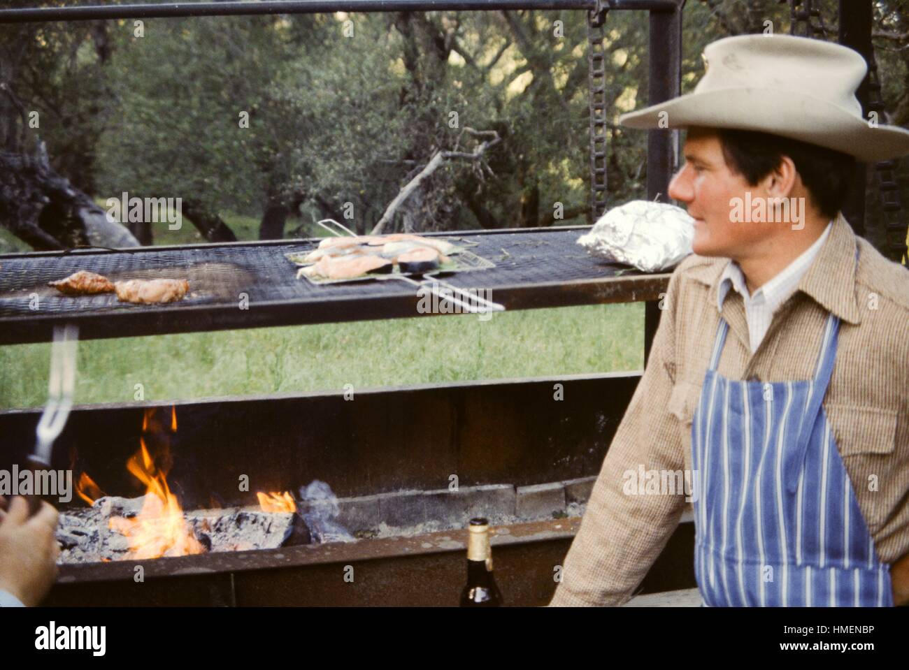 Man wearing a blue apron and cowboy hat grilling fish and meat over a large barbecue, 1975. Stock Photo
